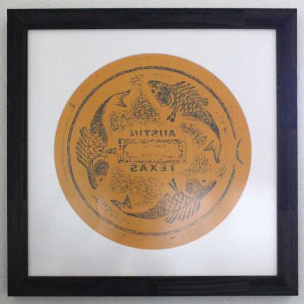 A blue and green rubbing on orange paper, in a circular mat, in a square dark purple frame. The rubbing design features three stylized fish chasing each other around a circle, with the words AUSTIN TEXAS mirror-reversed at the center.