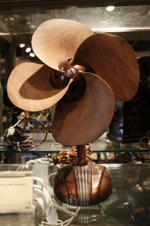 A desk fan, in front view, with no cage, and large free-spinning blades made of brown leather. The base is also brown and has attractive Art Deco styling elements.