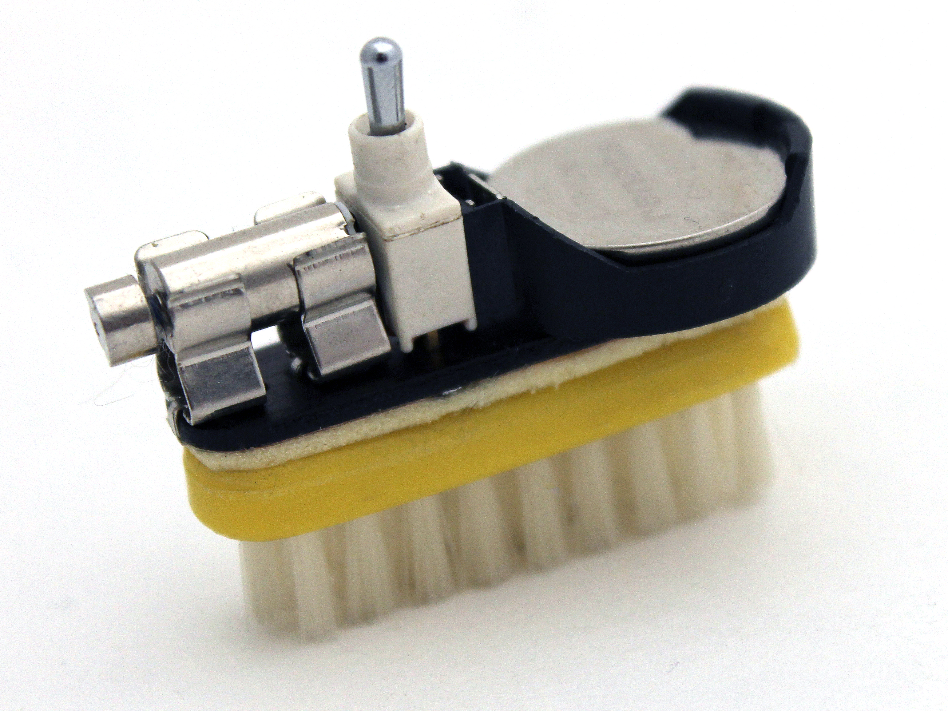 A yellow toothbrush head, with white bristles, that's been cut from the handle. The bristles are on the ground, and there's a layer of foam tape on the back of the head securing a small piece of black phenolic board that matches the shape of the head. On top of the phenolic board are a small pager motor, mounted in fuse clips, a toggle switch, and a CR2032 coin cell battery holder with a battery inside it.