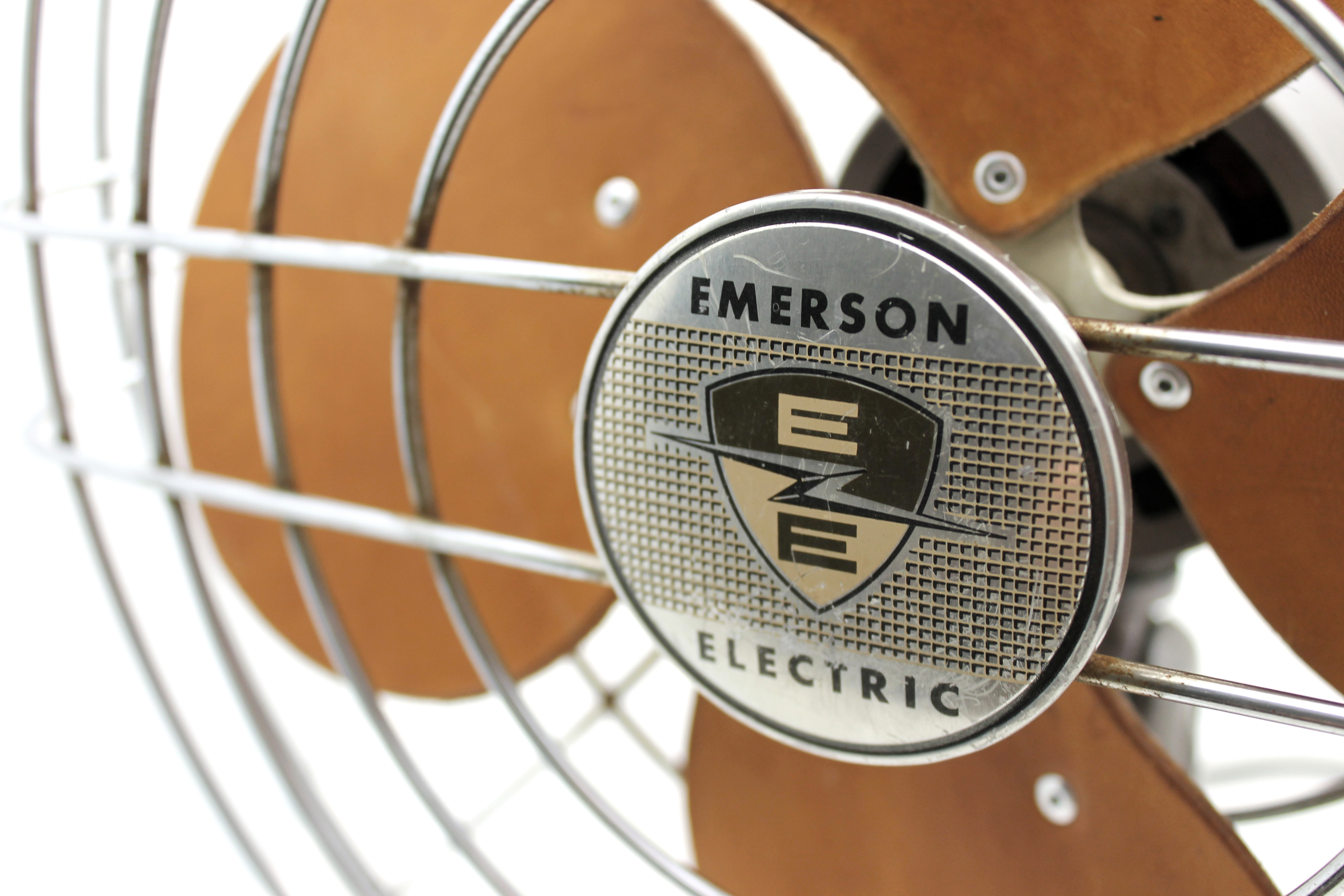 A close up shot of the EMERSON ELECTRIC logo medallion on the front of a wire fan cage, with the replaced leather blades out of focus behind.